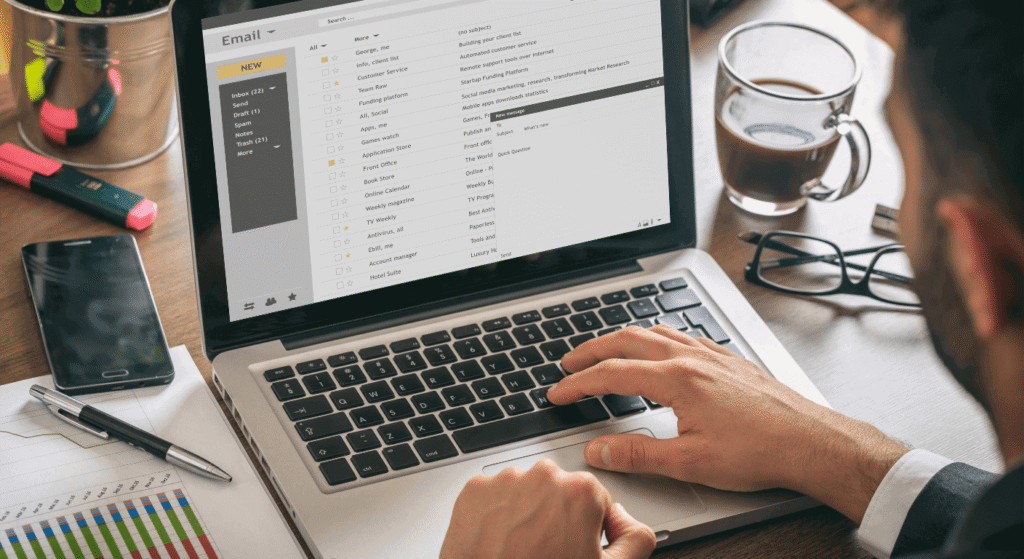 A person is checking their email on a laptop, surrounded by a coffee cup, smartphone, and paperwork with graphs.